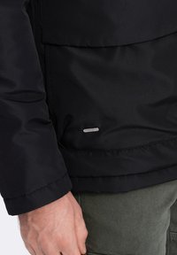 Černá zateplená bunda s hladkým povrchem, která obsahuje diskrétní kovovou nášivku a měkkou texturu. Viditelné švy na rukávech.