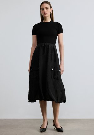 Naine seisab, kandes musta lühikeste varrukatega pasteldatud t-särki ja kortsulist musta midi-seelikut suured ees taskud ning mustad kontsakingad.