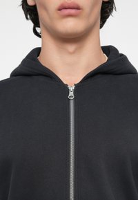 Melna kapuchekā ar rāvējslēdzēju, kas izgatavota no mīksta kokvilnas maisījuma, ar sudraba rāvējslēdzēja, kapuci un atbrīvotu piegriezumu ar gludu tekstūru.