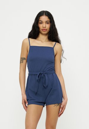 Kobieta o długich ciemnych włosach w niebieskim kombinezonie bez rękawów z paskiem do wiązania, ukazująca tatuaże na lewej ręce i prawej uda, stoi na białym tle.