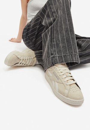 Bézs Reebok sportcipő textúrázott suede anyagból, perforált orr résszel és lapos fűzővel. Szürke csíkos, bőszárú nadrággal párosítva.