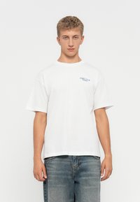 Hvit bomulls T-skjorte med korte ermer, med en blå trykt logo på venstre bryst, parret med falmede blå denimjeans.