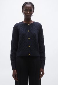 Ζακέτα πλεκτή με κάβελ χρώμα ναυτικού, με στρογγυλή λαιμόκοψη, έξι χρυσά κουμπιά στο μπροστινό μέρος και μακριά μανίκια. Κοντό σχέδιο.