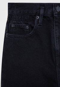 Gros plan sur la poche d'un jean en denim noir avec rivets, passants de ceinture, et fermeture par bouton en métal sur la ceinture.