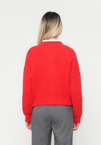 Suéter tricotado vermelho e fofinho, com decote redondo e ombros caídos, combinado com calças cinzentas com riscas. A vista de trás destaca o tecido texturizado.