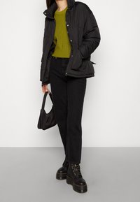 Schwarze Steppjacke aus strukturiertem Stoff, mit Knopfverschluss, kombiniert mit einem grünen gerippten Pullover, schwarzen geraden Jeans und schwarzen Plateaustiefeln.