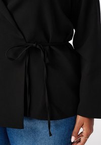 Zwarte wikkel top van gladde stof, met een striksluiting in de taille en lange mouwen. Gecombineerd met blauwe spijkerbroek.