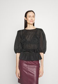 Musta pörhöhiha blouse, jossa on teksturoitu kuvio, kuminauhakiristys vyötäröllä ja röyhelöhelma, yhdistettynä burgundinväriseen nahkakuosiin.