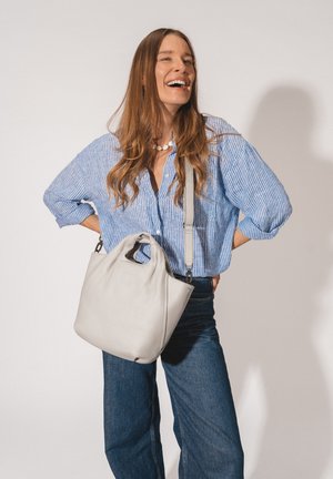 Lächelnde Frau mit langen Haaren trägt blau gestreiftes Hemd, Jeans, Muschel-Halskette und hält eine große hellgraue Ledertasche über der Schulter.