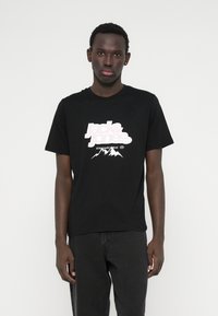 Μαύρη βαμβακερή μπλούζα με στρογγυλή λαιμόκοψη και κοντά μανίκια, διακοσμημένη με ένα λευκό γραφικό λογότυπο και σχέδιο βουνού μπροστά. Φορεμένη με σκούρα τζιν.