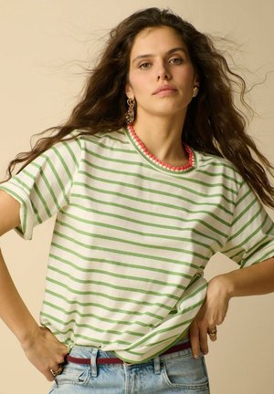 Femme aux longs cheveux ondulés porte une chemise rayée verte et blanche avec un bord rose, jean bleu, ceinture rouge, boucles d'oreilles et bagues en or, posant les mains sur les hanches.