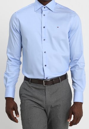 Mężczyzna w jasnoniebieskiej koszuli zapinanej na guziki, wpuszczonej w szare spodnie z czarnym paskiem, stojący na jednolitym tle.