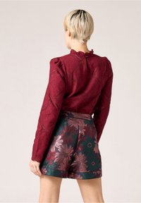 Burgundy tekstuuri blouse puff-kätistega ja rüüdega kaelusega, kombineeritud rohelise seeliku ja roosade ja kuldsete lillemusteritega.