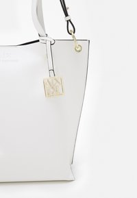 Bílá taška přes rameno s texturovaným povrchem, černými detaily, zlatým kováním a označením loga připevněným na ramenním popruhu.