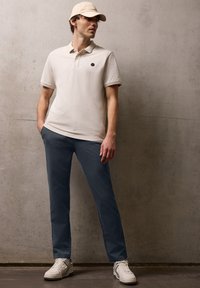 Mężczyzna w beżowej koszulce polo, pasującej czapce, niebieskich spodniach i białych trampkach stoi na tle gładkiej betonowej ściany.