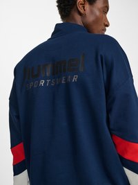 Person, die eine marineblaue Sportjacke mit roten und weißen Ärmelstreifen trägt und auf deren Rücken "hummel SPORTSWEAR" aufgedruckt ist.