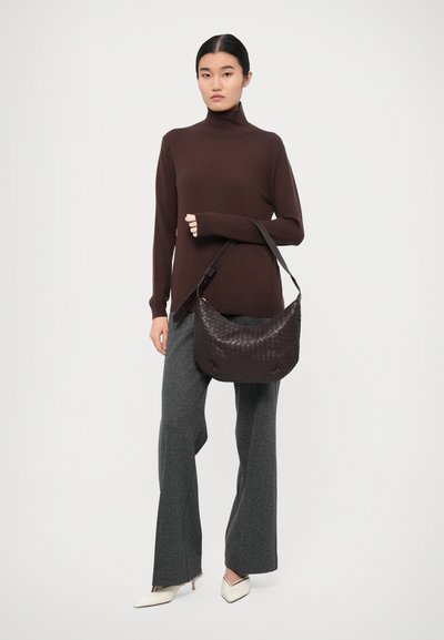 Borsa a tracolla in pelle intrecciata marrone scuro, abbinata a una maglietta marrone e pantaloni grigi a campana. Tacchi a punta bianchi.