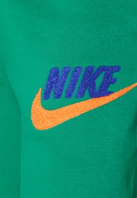 Grön sweatshirt-tyg med en texturerad blå "NIKE"-logotyp och ett klart orange Swoosh-emblem, båda tydligt synliga.