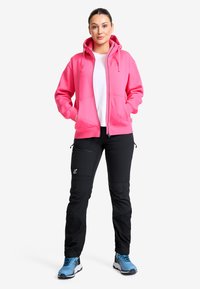 Rosa zip-up hoodie med en stor huva och framfickor, kombinerad med svarta byxor med dragkedjor och blå sportskor.