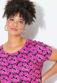 Camisa rosa de manga corta con un patrón de corazones sobre un fondo negro, confeccionada en tejido suave. Cuello redondeado y corte relajado.