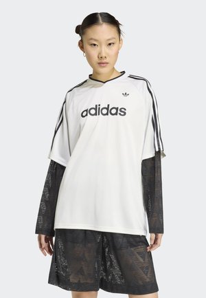 Donna che indossa una maglietta bianca oversize Adidas con strisce nere e logo, sovrapposta a un top trasparente nero con motivi e pantaloncini.