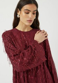 Viininpunainen mekko, jossa pystysuuntaiset paljettirivit ja hapsukoristelu. Pitkät hihat, teksturoitu kangas, pyöreä pääntie ja rento istuvuus.