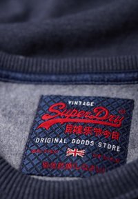 Etichetta in tessuto blu con testo rosso su uno sfondo testurizzato, con "Super Dry" in evidenza. L'etichetta include una bandiera britannica e l'etichetta "Vintage".