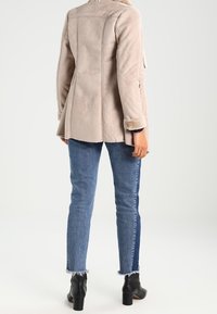 Světlá béžová bunda z umělé semiše s rozšířeným střihem, viditelnými švy a plyšovými rukávy. Kombinována s tmavě modrými džíny a černými kotníkovými botami.