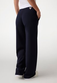 Marineblaue weite Hosen mit einem weißen Bund, seitlichen Streifen und einem dreieckigen Logo-Patch. Hergestellt aus weichem Stoff mit glatter Textur.