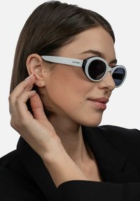 Aurinkolasit, joissa on pyöreä muoto, mustat linssit ja valkoinen kehys. Suunnittelussa on rohkea, kontrastinen väriteema.