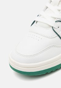 Zapatilla blanca con parte superior de sintético texturizado, suela de goma verde, puntera reforzada y detalles perforados en el empeine.