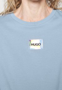 Světlomodrá košile s iridescentním čtvercovým štítkem, na kterém je ve středu na hrudi pod výstřihem umístěno slovo "HUGO".