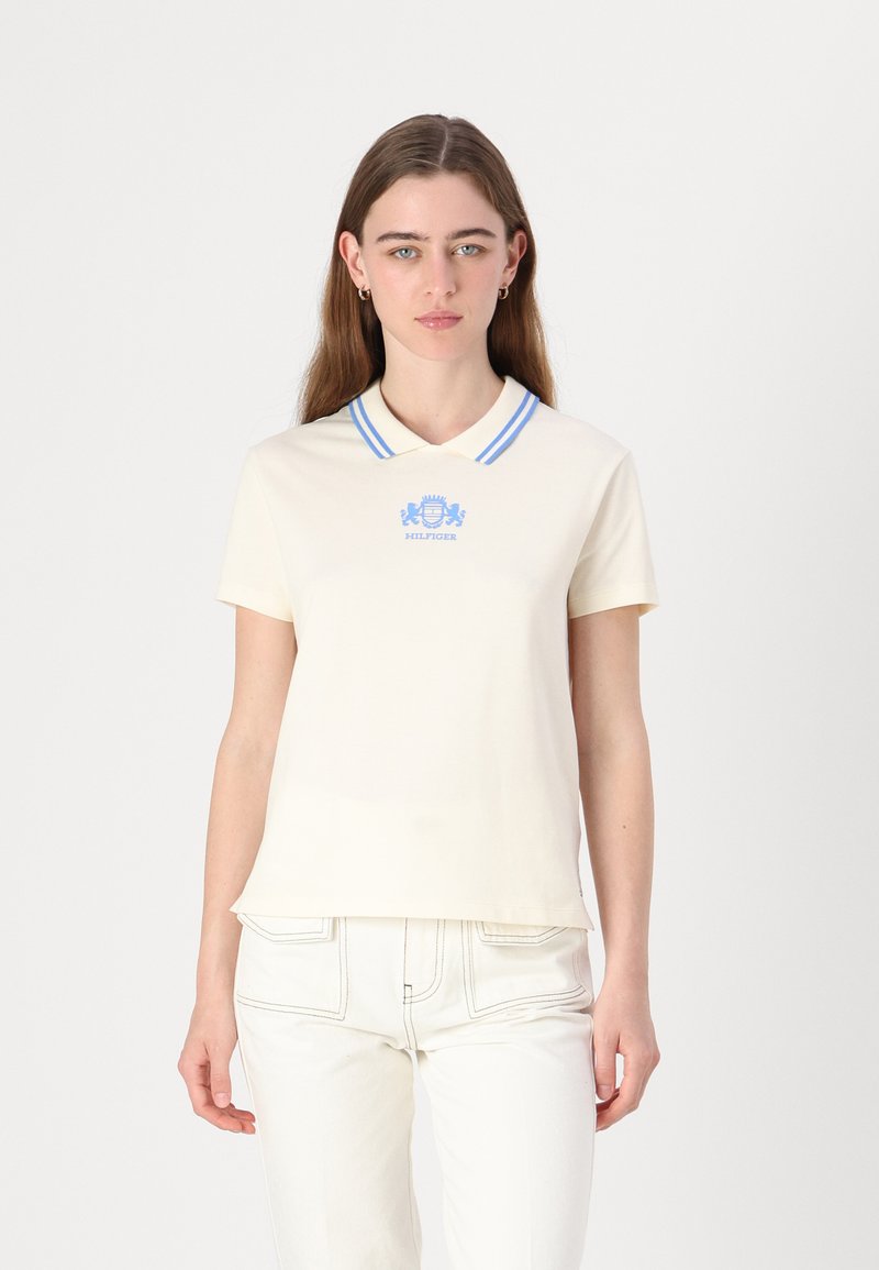 Polo shirt à manches courtes crème avec un col rayé bleu et un logo brodé sur la poitrine. Porté avec un pantalon blanc, texture de tissu lisse.