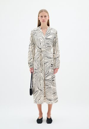 Langes, cremefarbenes Kleid mit schwarzen, geschwungenen Linien, einem V-Ausschnitt, tailliertem Bund und langen Ärmeln. Kombiniert mit schwarzen, hohen Schuhen und einer kleinen Tasche.