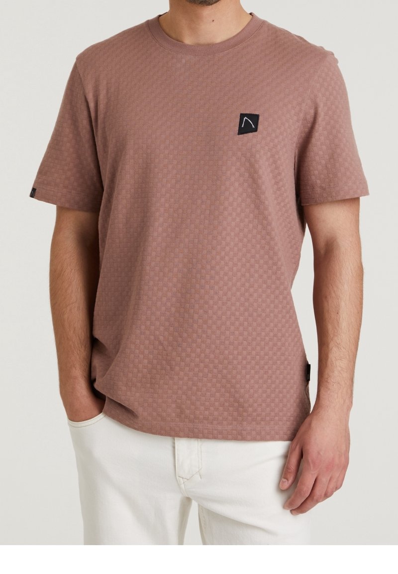 Mężczyzna w szorstkiej, różowej koszulce o krótkim rękawie z małą czarną naszywką na piersi oraz białych spodniach. Ręce w luzie.