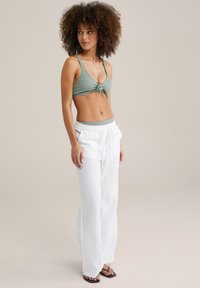 Žalias rauktas bikinio viršus su mazgo detale, derinamas su baltais laisvais lino kelnėmis ir juodomis plokščiomis šlepetėmis. Neutralus fonas.