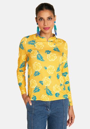 Langärmeliges gelbes Oberteil mit blauem Blumenmuster, Kragen, weicher Textur und tailliertem Design. Kombiniert mit blauen Jeans.