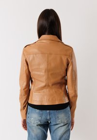 Giacca di pelle tan con vestibilità su misura, spalline e dettagli in cucitura visibili. Orlo nero a coste, indossata sopra jeans chiari scoloriti.
