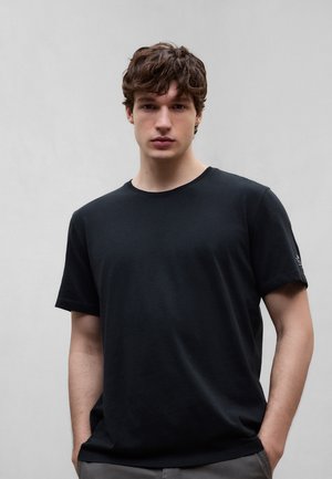 Ung mand med krøllet brunt hår iført en simpel sort t-shirt med korte ærmer, stående foran en neutral grå baggrund.