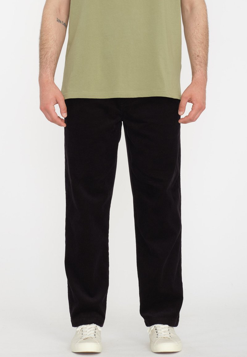 Pantalon en velours côtelé noir à jambe droite, présentant des côtes verticales et une texture douce. Associé à un t-shirt vert clair et des baskets blanches.