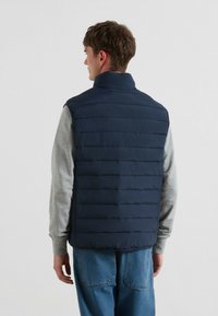 Marineblaues, gepolstertes Westchen mit Stehkragen und horizontalen Nähten. Getragen über einem hellgrauen Langarmshirt und blauen Jeans.