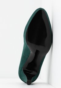 Chaussure à talon haut en suède vert à bout pointu avec semelle noire lisse et brillante, posée verticalement contre un mur blanc et sur un fond blanc.