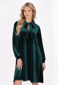 Rochie verde din catifea, cu mâneci lungi, închidere cu cravată la decolteu și croială lejeră. Textură moale cu o suprafață netedă și lucioasă.