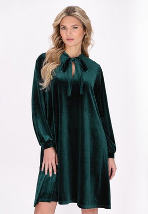 Rochie verde din catifea, cu mâneci lungi, închidere cu cravată la decolteu și croială lejeră. Textură moale cu o suprafață netedă și lucioasă.