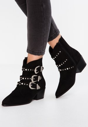 Bottines en daim noir avec trois boucles argentées et clous, portées avec un jean gris foncé raccourci sur fond blanc.