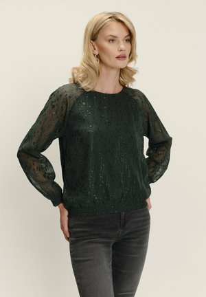 Blondynka w przezroczystej, ciemnozielonej bluzce w błyszczący kropkowany wzór oraz czarnych dżinsach, stojąca z rękami na biodrach na jednolitym tle.