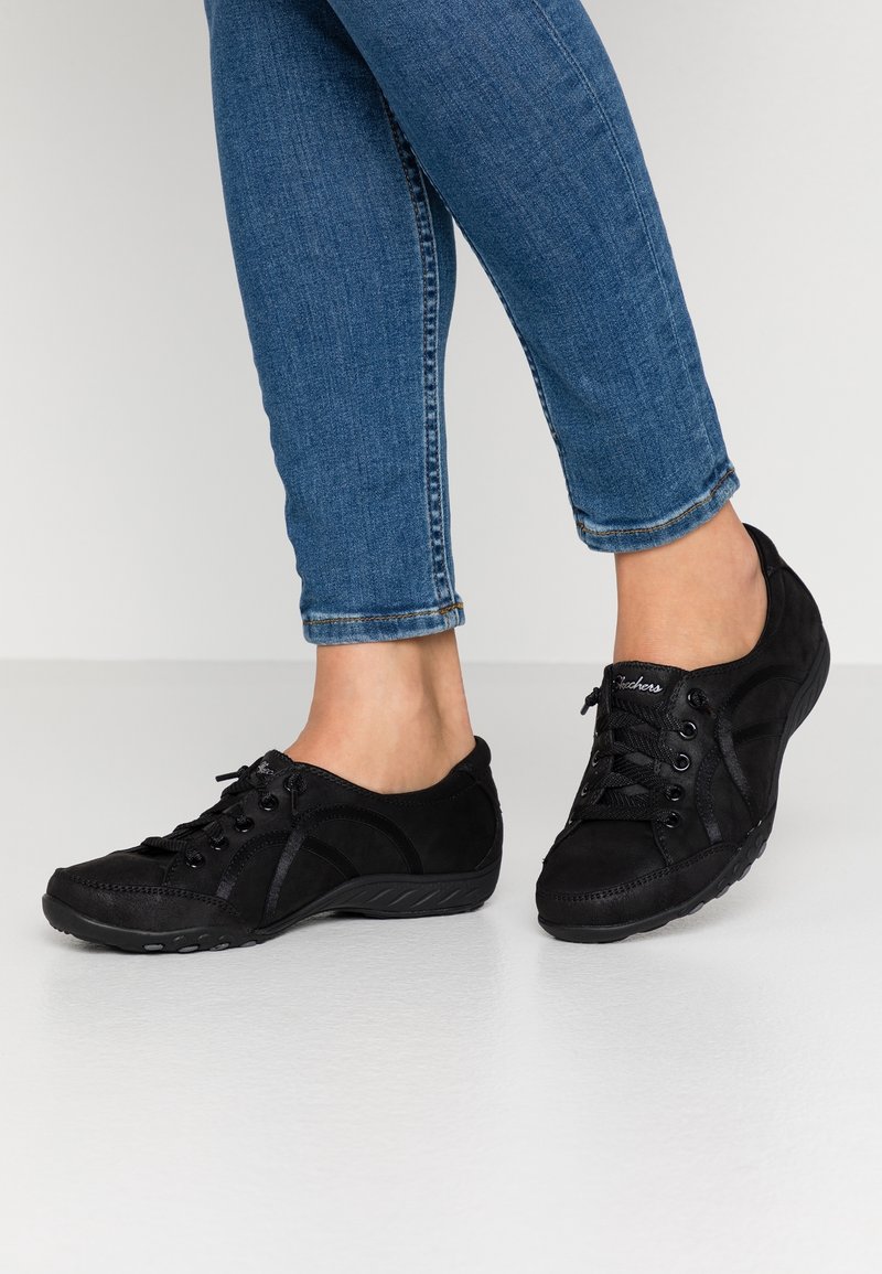 Personne portant des chaussures décontractées noires à lacets et un jean skinny bleu debout sur un sol blanc.