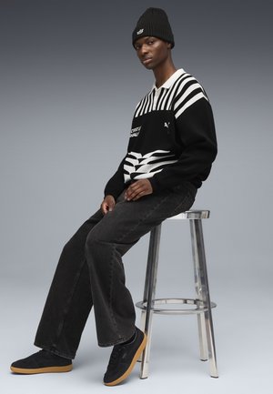 Zwarte trui met witte strepen, logo en geometrisch patroon; donkere jeans; zwarte sneakers met gumrubberen zool; zittend op een zilveren kruk.