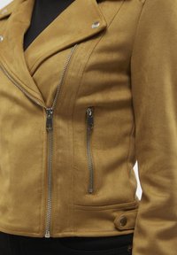 Bruine motorjack van neppersuède met zilveren ritsen, ritszakken en drukknopdetails op de kraag en zoom. Getailleerd silhouet.