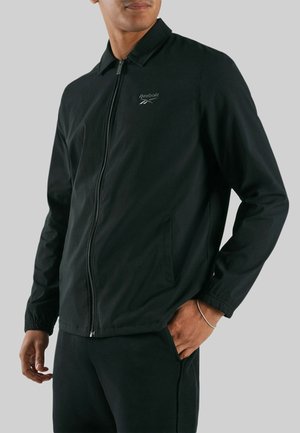 Man in een zwarte Reebok-jas met ritssluiting, kraag en elastische manchetten, staand met één hand in de zak tegen een effen achtergrond.